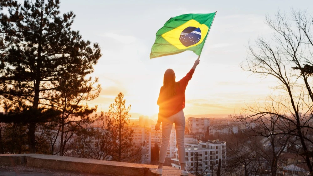 A woman holding a Brazilian flag at sunset on a mountain overlooking city buildings, symbolising Brazil gambling regulations and the country’s evolving landscape.