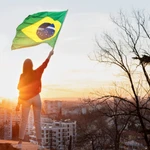 A woman holding a Brazilian flag at sunset on a mountain overlooking city buildings, symbolising Brazil gambling regulations and the country’s evolving landscape.